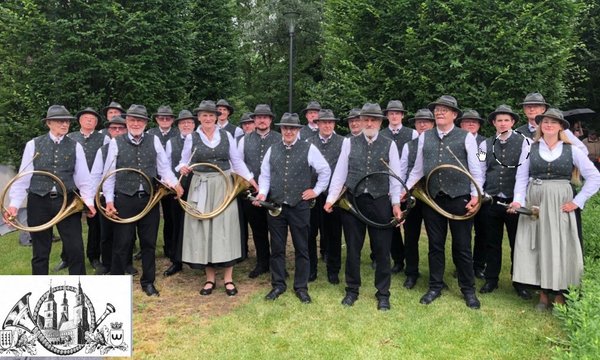 Eine Gruppe von Personen trägt Trachten und hält Jagdhörner, steht lächelnd in einem grünen Garten. Das Bild zeigt ein Logo mit Musikinstrumenten und einer Stadtansicht.