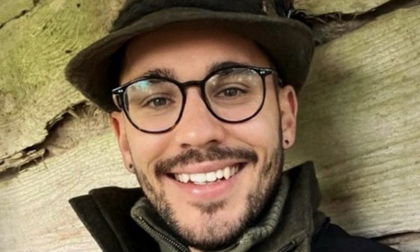 Ein Mann lächelt, trägt eine Brille und einen Hut, sitzt in einer grünen Jacke mit Logo „PS“, vor einer hölzernen Wand.